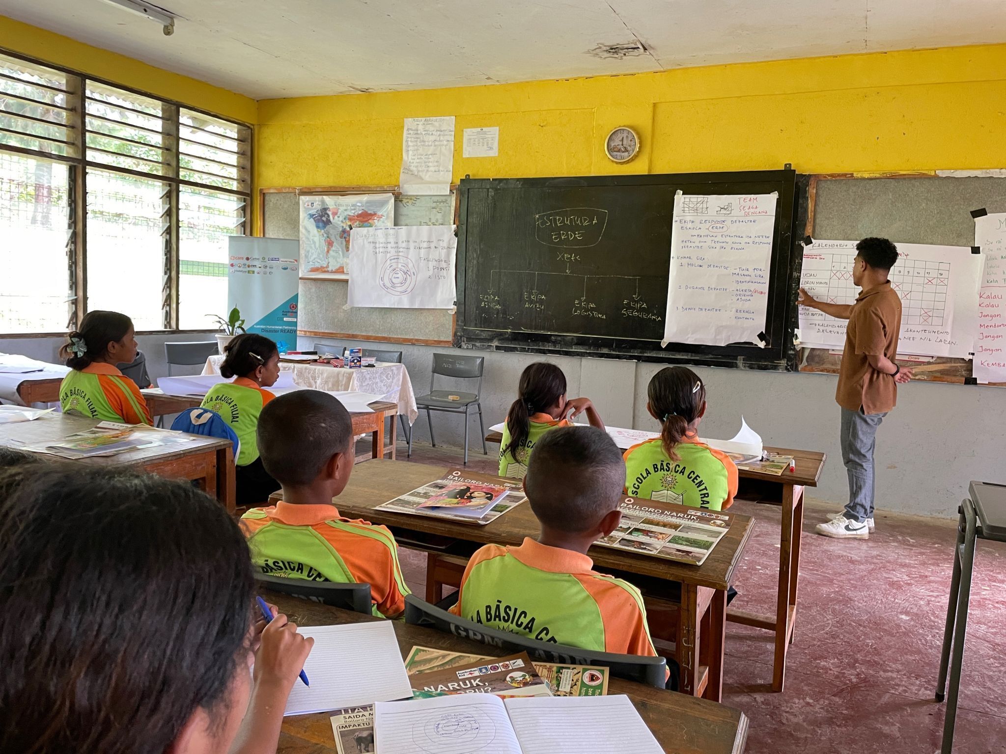 A young leader teaches environmental resilience strategies to students in a classroom, pointing to a blackboard.